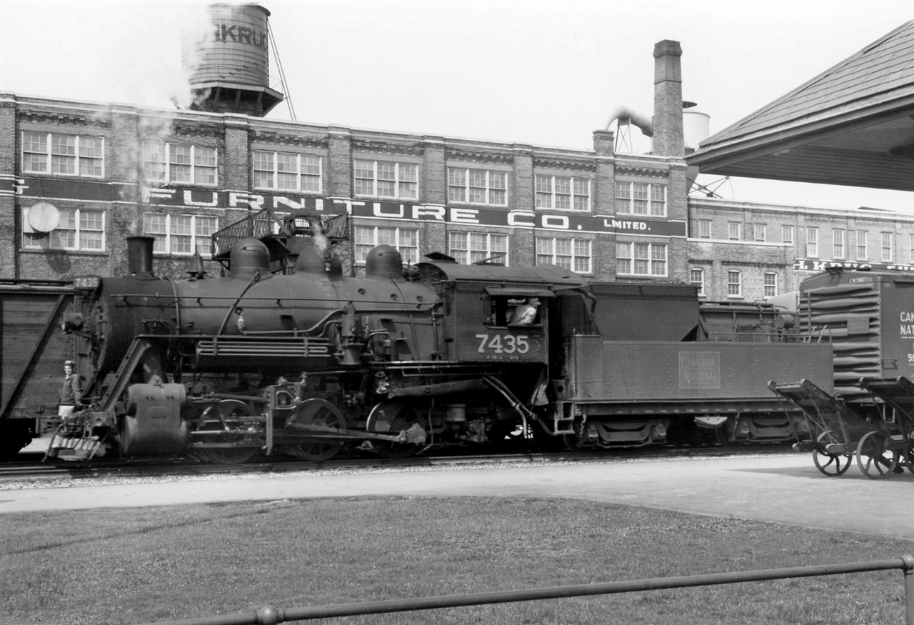 Canadian National 0-6-0 7435 switches at Kitchener in front of the station in 1958, with one of the crew riding the front pilot as another looks back on their train. Baggage carts for servicing passenger trains are posed by the station under the awning. The Krug Furniture Company building, still standing today, is visible in the background along with its water tower.