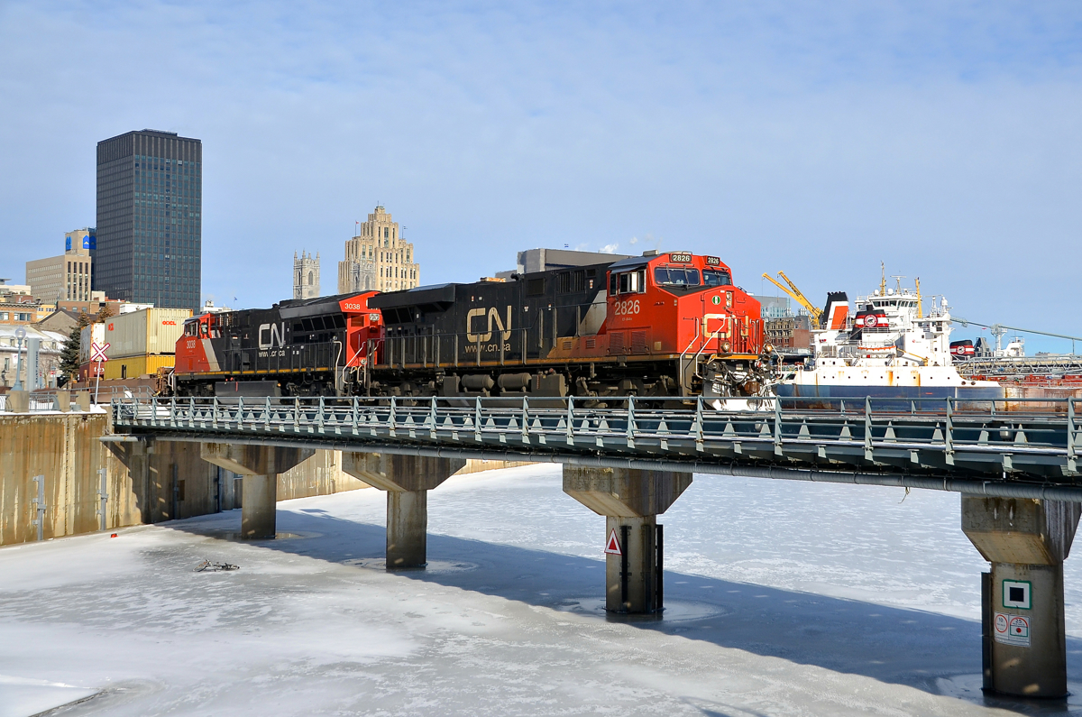 CN 149 and 3 ship funnels. CN 2826 & CN 3038 lead CN 149 over the frozen entrance to the Lachine canal on a cold but bright morning. In the distance the funnels of three ships are visible, laying over for the winter in Montreal. From closest to furthest, they are the Tim S. Dool, the Baie St-Paul and the Algoma Navigator.