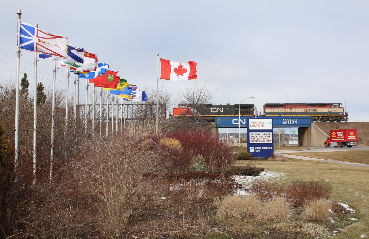 CN train 384 with a colourful BC Rail leader passes the colourful arrangement of provincial flags at Chris Hadfield park in Milton.
