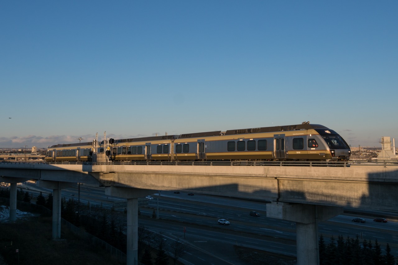 It's 17:09 on the coldest day of the winter yet and Union Pearson Express 4091 is knocking down the advance to Pearson on the west track of the Pearson Sub. 1010, 3004 and 1002 will make 6 more roundtrips before returning to either Don or Willowbrook Yard or the TMC, while the passengers onboard may find themselves thousands of miles away in warmer weather by the time that happens. Shadowed by the guideway and value park garage, traffic comes and goes. By the time this consist returns to the airport 75 minutes later, it is possible that passengers from the landing plane in the background will have made it through customs and baggage claim, ready to board the UP Express for a trip into the city.