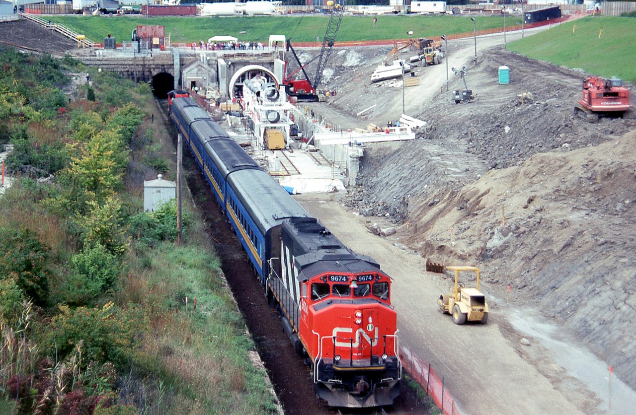 Canadian National's St. Clair Tunnel at Sarnia was too small to handle higher cars such as double stacks, so the decision was made to bore a new, larger tunnel. A special ceremony was held on September 16th 1993 to celebrate the start of the new bore. The old tunnel was built in 1891, 6025 feet long and had a bore diameter 19 feet 10 inches. The new tunnel would be opened in 1994, 6129 feet long and have a bore diameter of 27 feet 6 inches.

Here is a special passenger train that took special guests through the old tunnel to Port Huron, where there was an elaborate lunch before the return trip. There was also a ceremonial function in a large tent in Sarnia.

In the photo we see the passenger special, made up of two CN GP40-2W's bracketing 5 VIA passenger cars, starting through the old tunnel on the left. The green paint showing on the bell of CN 9674 gives off her former GO Transit heritage (one of 10 purchased from GO a few years back). On the right the Lovat TBM (tunnel boring machine) is ready to start the new bore.