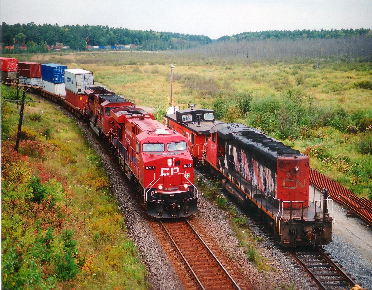 Shared assets in the Shared Asset Zone.  CP #102 with 8705 and 9113 snakes southward past CN 5257 & Van 79707 waiting it out on the MoW siding. View long-lensed from Hwy 69 overpass. Medium format 120- 400 ISO film for anyone interested. From near Parry Sound to just south of Sudbury CN and CP share trackage. CN line is used for southbounds, CP track for northbounds.