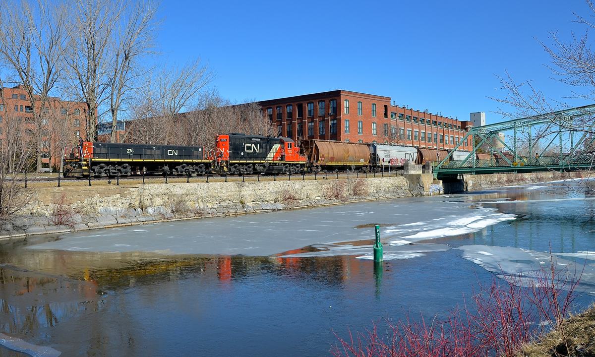 Along the Lachine canal. Slug CN 274 and GP9 CN 7272 have left the Robin Hood mill with 7 empties as they head west along East Side Canal Bank Spur. Once a common sight along both sides of the Lachine Canal, trains are now very rare, with Robin Hood mill being the only rail-served client along the length of the entire canal. The train has just crossed over Charlevoix street, which the crew had to flag.
