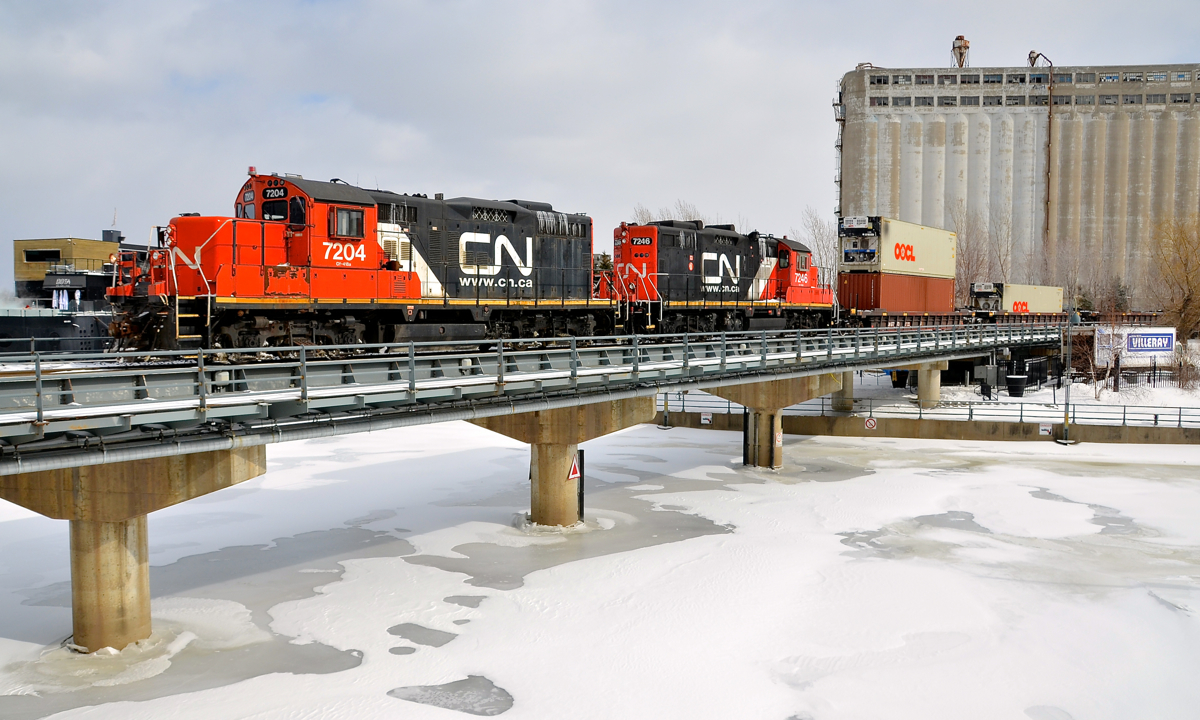 Double GP9's and double stacks into the Port of Montreal. A CN transfer enters the Port of Montreal with a pair of GP9's (CN 7204 & CN 7246, both very clean and in the current CN paint scheme) with a long string of well cars, with double stacks, single stacks and some empties. The train is crossing over the eastern end of the Lachine canal and passing the eastern edge of grain elevator #5.