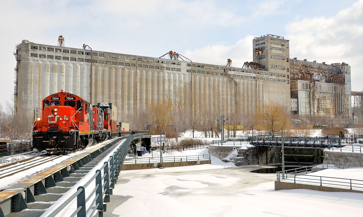 Stopped with grain elevator #5 behind. A CN transfer is about to enter the Port of Montreal with a pair of GP9's (CN 7204 & CN 7246, both very clean and in the current CN paint scheme) with a long string of well cars, with double stacks, single stacks and some empties. The train is stopped over the eastern end of the Lachine canal, awaiting port security and is passing grain elevator #5.