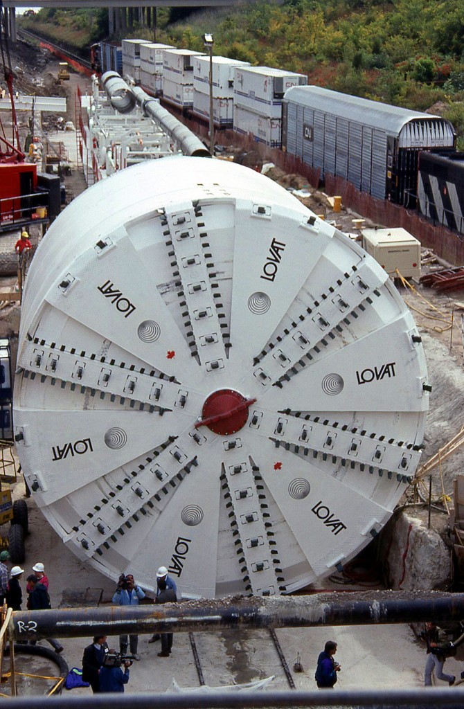 Canadian National's St. Clair Tunnel at Sarnia was too small to handle higher cars such as Double stacks so the decision was made to bore a new, larger tunnel. A special ceremony was held on Sept 16, 1993 to celebrate the start of the new bore. The old tunnel was built in 1891, 6025 ft long and had a bore diameter 19 ft 10 inches. The new tunnel would be opened in 1994 ,6129 ft long and have a bore diameter of 27 ft 6 inches.

Seen here is "Excalibore", the name of the tunnel boring machine (TBM) made by the Lovat Tunnel Manufacturing Co., ready to start on the new larger tunnel on the Canadian side. In the background, a special CN train of high-clearance cars including double stacks, an autorack and a hi-cube auto parts boxcar, sandwiched by a pair of CN GP40-2W's, sit on the tracks nearby that lead through the old St. Clair tunnel.