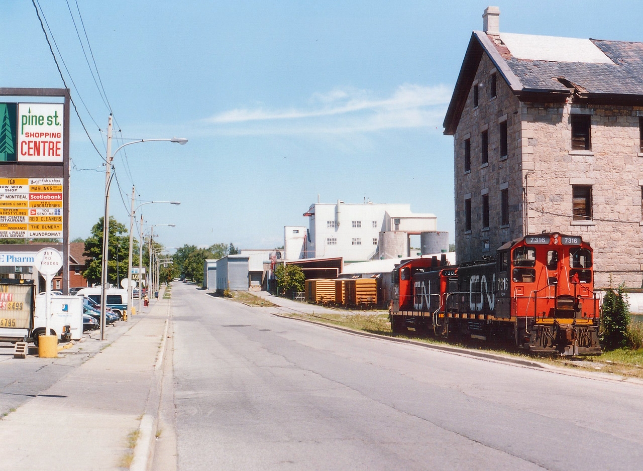 This is a once common scene that has almost vanished in this country. The street running.  CN 7305 and 7316 have just run down the center of Pine St from the CN Canal Line and are seen following what was once NS&T trackage to the east side of the street to service the Gallagher Paper company, located just north of Albert St W.on Pine. Upon return the local took out 3 of the yellow boxcars as seen. Gallagher was served two days a week, but not long after Trillium took over this run from CN that year, Gallagher shut down and that was the end of the Pine Street Running. I do not recall Trillium actually servicing this plant. All track visible in this photo has been removed, the old stone building on the right has been restored, and the Pine St plaza has been updated.