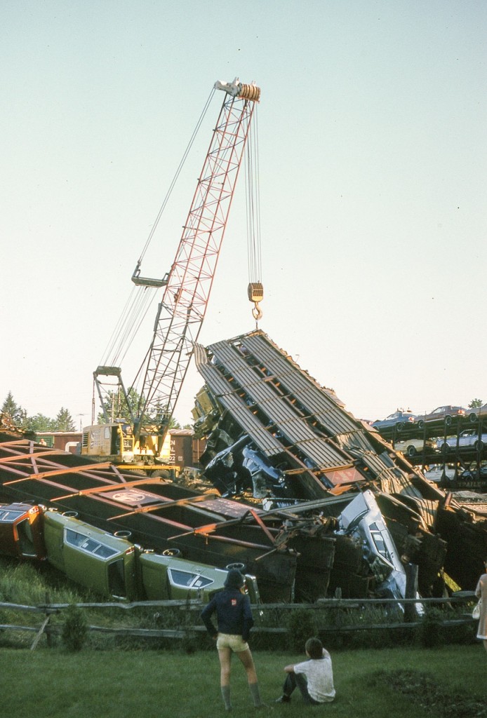 Witnessing probably the biggest train derailment that ever happened in Simcoe, a couple of spectators watch the cleanup from the backyard of their home. Eastbound N&W train, loaded with new autos, went off the tracks, with lead unit going off the overpass at Norfolk St., resulting in the deaths of two crew members. Damage was over a million dollars, and lets not forget these are 1975 dollars. Priority for the cleanup crew was getting the line open, and in public areas the crowds were entertained watching rail cars getting thrown around, and my take was that this incident saved a lot of people from owning Pintos. There were two private driveways in which I could shoot from; sadly enough, the next door drive featured a guy leaning on his new Pinto wagon watching all those others get thrown around like they were worthless. One could only imagine what was going thru his mind. I elected to submit this photo because, well, the scenery was better.