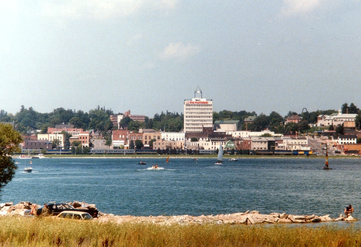As seen across Kempenfeldt Bay, an arm of Lake Simcoe, the VIA Canadian #9 heads northward past downtown Barrie. Power is VIA 6516, 6621 and 6618. The train has just left the station stop at Allandale, and for photography, everywhere is into the morning sun save for this location, as far as I knew. Times have changed. The track has been replaced with a bicycle/hiking trail, after the train service was discontinued in 1997 and the rails lifted. About center in this image stood the early Barrie station, it was unfortunately demolished back in 1962.