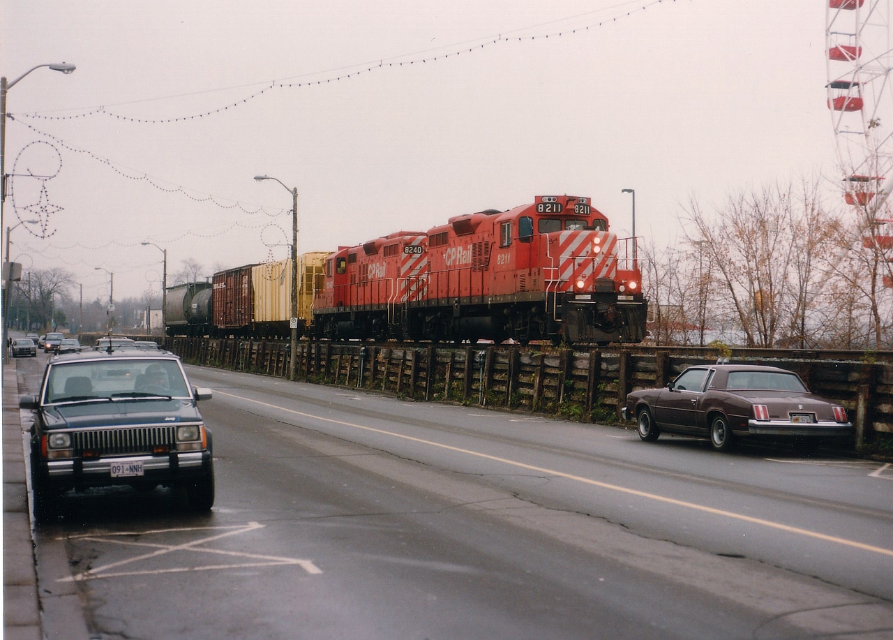 For a change of pace, here is an image taken in downtown Niagara Falls' Clifton Hill tourist area of a train before it reaches Clifton on its way west.........this is obviously not very photogenic as far as locations go, but it is interesting to see just how narrow the right-of-way is along here. Not exactly world-class touristy to park your car and have a train literally looming overhead !!! And some of those trains could be intimidating in size and especially noise.  Not this one though. It is off season, thus quiet in the neighbourhood; CP 8211 and 8240 on their way back to Welland after interchanging with CN by way of the industrial connector that used to run along Park Av and right thru the intersection of Victoria Av and Bridge St. No wonder the city was so happy to get CP out of town...... For some reason I cannot recall, I was chauffeured around on this day, as the Jeep parked on the left has Peter Hoople, railfan/chauffeur driver, waiting as I grab this quick shot. (I sure as heck didn't pay him.)