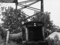 CN 1211 trundles back into town after working the Esso depot that used to occupy the south side of the harbour just to the west of the CP Parry Sound trestle.  The huge black storage tanks are long gone, replaced by modern boat marina, and with it went all the industrial trackage in Parry Sound. This view shows a string of tank cars, and the CP trestle is above it. Rough old photo, but this is one location where at absolutely no time of the day was the sun in a favourable position. So dull days were needed. The track connected with the CN mainline at mile 149.2, and also with the CP main, at the north edge of town. One could call this an early effort at shared assets, as CN used it at certain hours and CP other hours, with CN permission. When not in use this switcher was stationed along Great North Rd about 200 ft behind me, this side of Bowes St. What I do not know, however, is what year the local service ended and the track was pulled.