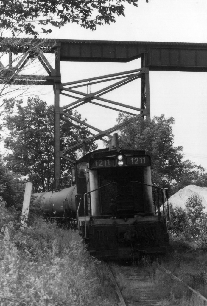 CN 1211 trundles back into town after working the Esso depot that used to occupy the south side of the harbour just to the west of the CP Parry Sound trestle.  The huge black storage tanks are long gone, replaced by modern boat marina, and with it went all the industrial trackage in Parry Sound. This view shows a string of tank cars, and the CP trestle is above it. Rough old photo, but this is one location where at absolutely no time of the day was the sun in a favourable position. So dull days were needed. The track connected with the CN mainline at mile 149.2, and also with the CP main, at the north edge of town. One could call this an early effort at shared assets, as CN used it at certain hours and CP other hours, with CN permission. When not in use this switcher was stationed along Great North Rd about 200 ft behind me, this side of Bowes St. What I do not know, however, is what year the local service ended and the track was pulled.