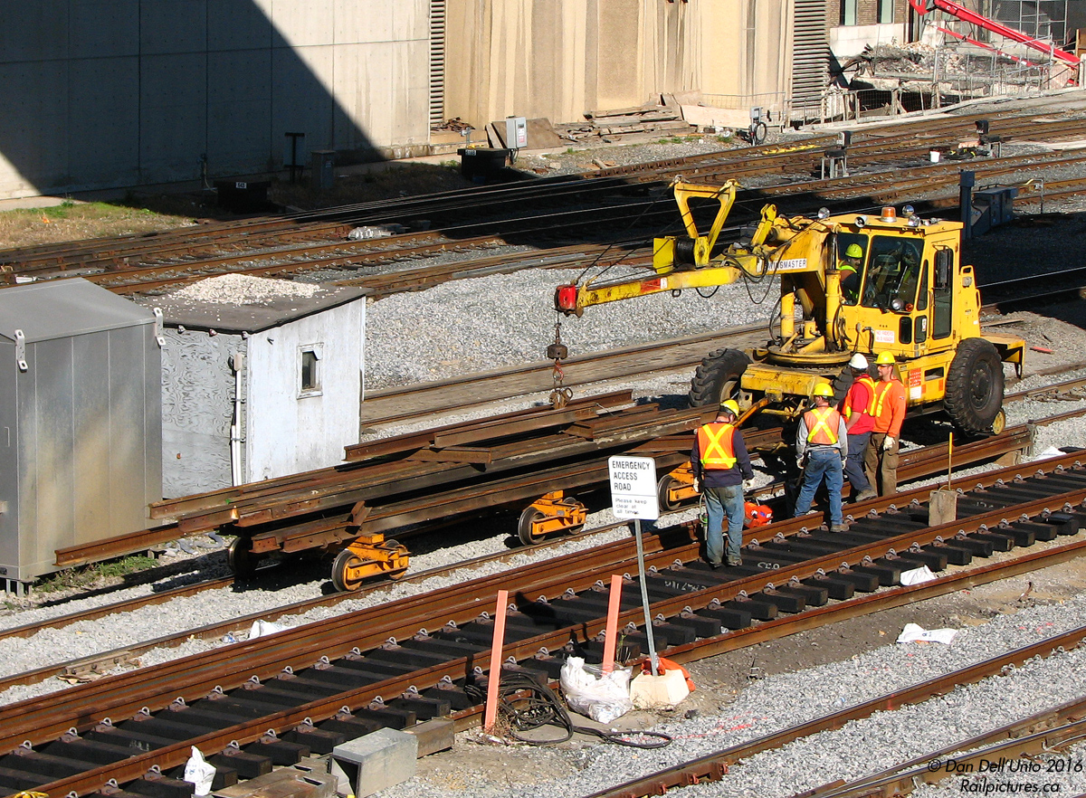 Lazy afternoon going-ons down at the station: a gang of Toronto Terminals Railway (TTR) employess work on new a track panel for replacing a section of trackage around Union Station, while their Swingmaster Swingloader passes by towing a load of old rail on bogies. Old wooden signal bungalows and modern galvanized steel ones mingle freely nearby, and new ballast is spread on neighbouring tracks that have recently been attended to. In the upper right, work continues on the Simcoe Street underpass project that will open to traffic in the future (you can see the remains of the old MofW storage building at the end of the Skywalk that was demolished).