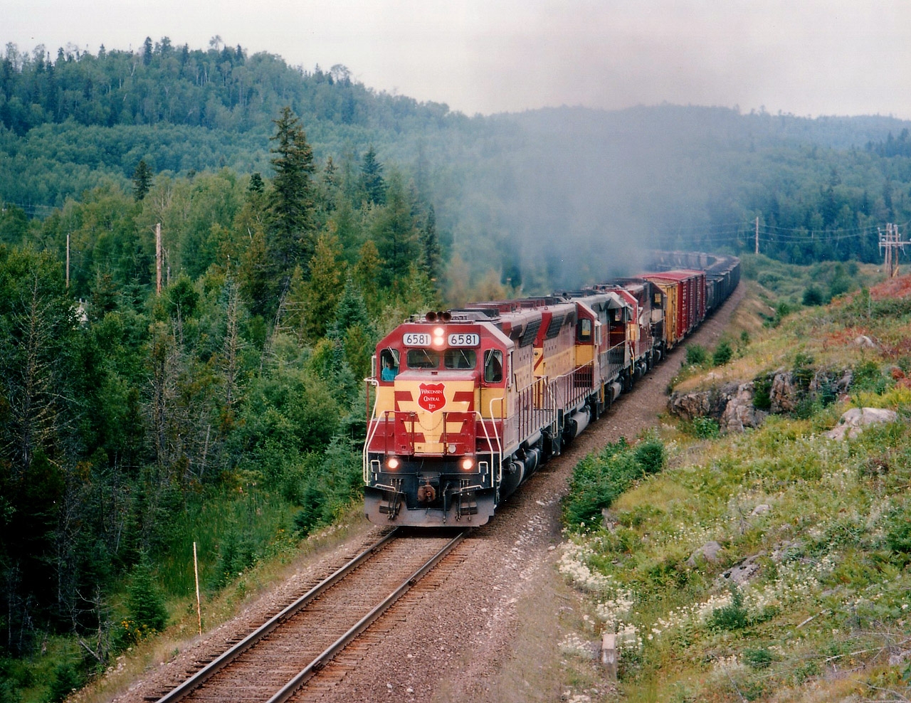 The daily #10 roars out of Hawk Junction on its late afternoon departure for Sault Ste. Marie. Five units on the head end for the climb. WC 6581, 6522, 6006, 6583 and 589. The 6006 is still in AC paint with its AC number, but WC heralds on the ends. Last unit 589 is a relatively scarce EMD SDL39, it is still active, but now down in Chile. In this photo it was just mooching a ride.