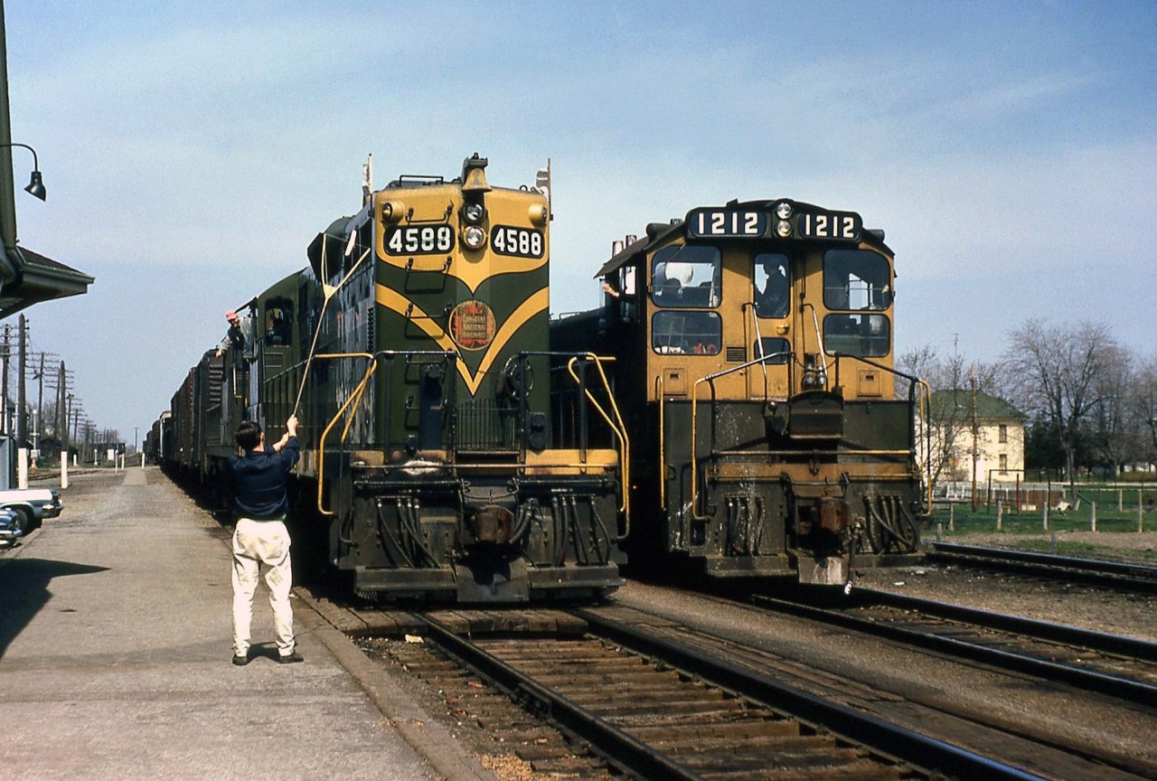 The Canadian National station operator at Glencoe stands on the platform with his train order hoop, about to pass orders to a westbound freight behind GP9 4588 in May 1968. SW1200RS 1212 waits on the passing track at the station, its crew giving a friendly wave out the cab window.