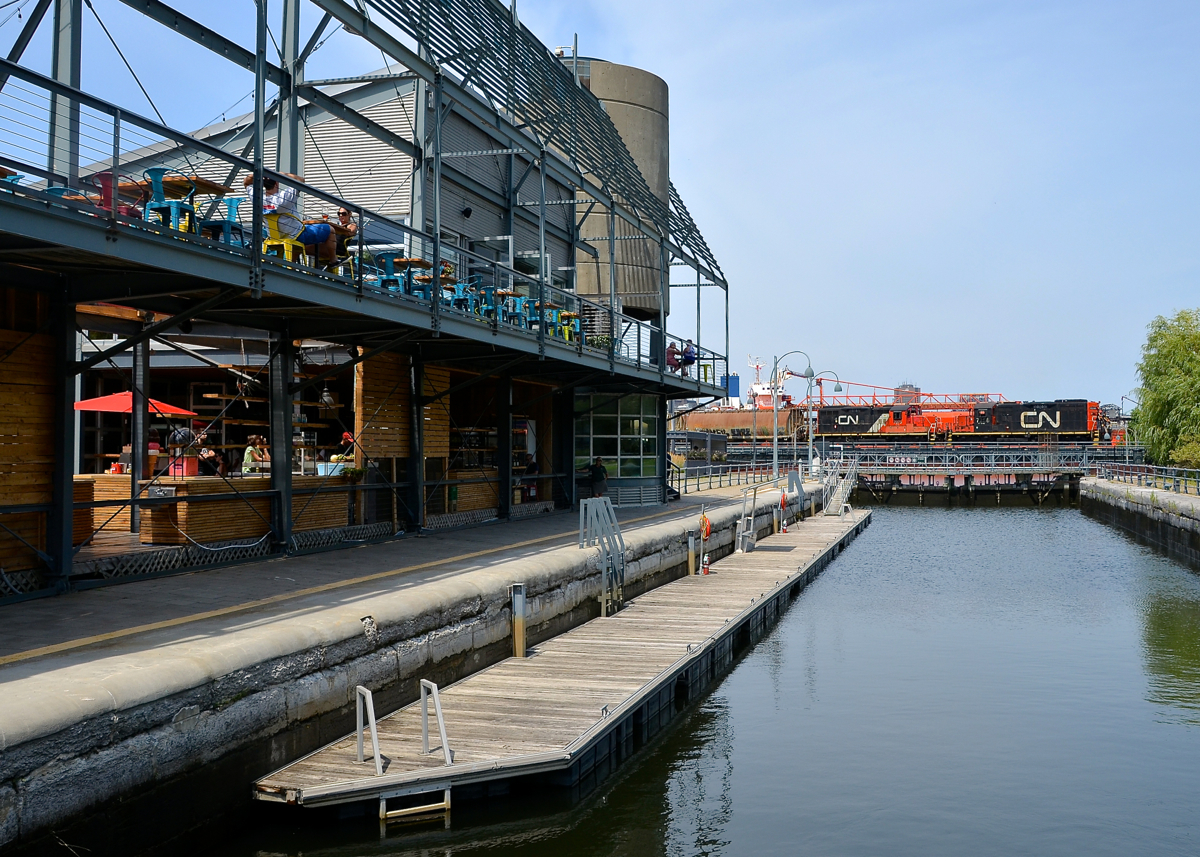 Over the lock and past the market. After coming into the Port of Montreal light, GP9's CN 7256 & CN 7075 head back with a short train. They are crossing over the eastern end of the Lachine Canal, where a lock exists for pleasure boats and passing the Éclusiers market at left.