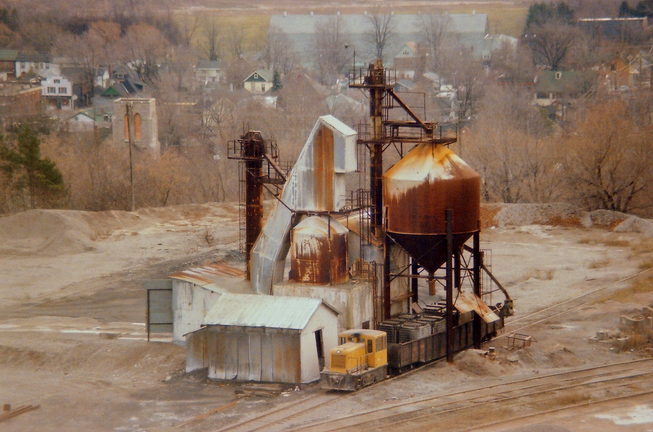 This image should appeal to those interested in the local history around Dundas. Currently, if you look over the side down into the valley town of Dundas from up on the Peak, you will see a modern upper-scale subdivision just below the CN mainline approximately Mile 4, Dundas Sub. This image predates all of that. The area once was the location south side of the tracks for quarry operations, limestone & dolomite/dolostone mostly; and the image is of the former Dolomite Refractories Ltd operation that existed there from 1945 to early 1977, after which Steetley took it over. The view shows the company switcher with a gondola of containers being loaded with dolomite. Perhaps this is destined for Dofasco? (Dolomite can be used as a flux for the smelting of Iron & Steel)Perhaps it is being shipped out to a marketer of potting soils or the like; dolomite is also used as an acid neutralizer. Many uses. The making of cement is another. This area also stockpiled crushed stone from the limestone quarry above (Canada Crushed Stone) which was used mainly in construction materials. Out of sight is rail connection to and from CN as well as a connection to the TH&B which at one time ran thru the valley joining the current trackage at Aberdeen Yd in Hamilton.  All this is gone, and looking around today, one would not realize it ever existed. Comments/corrections appreciated.