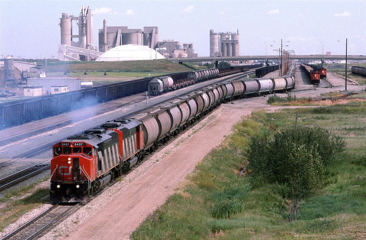 We have lots of interesting things in this photo. There is a loaded potash train heading west to North Vancouver. Potash was loaded near Watrous of Melville SK.
In the way of static trains, from left to right, there is an empty coal train (When I blow up the image, I think that I see a caboose on the far end), a strange assortment of cars in the next track. The car on the end appears to be a limestone car for Inland Cement seen in the background. Other cars in that line might be potash?, but the flats puzzle me - perhaps ribbon rail cars?.
There is another coal train, then a sulphur train and finally, I think that I see a wood product train (#359?)