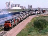 We have lots of interesting things in this photo. There is a loaded potash train heading west to North Vancouver. Potash was loaded near Watrous of Melville SK.
In the way of static trains, from left to right, there is an empty coal train (When I blow up the image, I think that I see a caboose on the far end), a strange assortment of cars in the next track. The car on the end appears to be a limestone car for Inland Cement seen in the background. Other cars in that line might be potash?, but the flats puzzle me - perhaps ribbon rail cars?.
There is another coal train, then a sulphur train and finally, I think that I see a wood product train (#359?)