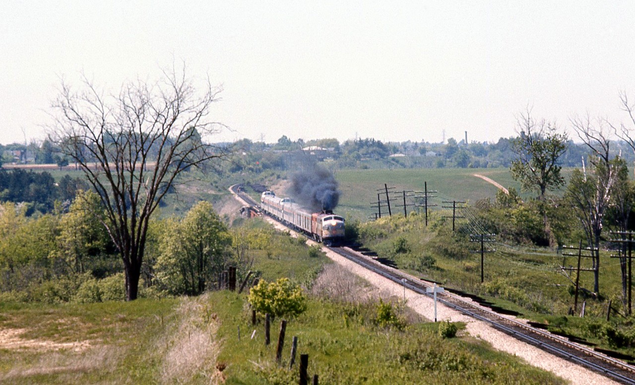 Under a cloud of MLW smoke from the trailing FPA2, The Canadian (#12) heads south after passing through Woodbridge, crossing over the Humber River bridge on Canadian Pacific's MacTier Sub. The train is approaching the white "Emery" mileboard sign, and the CN Halton Sub overpass northwest of Islington and Steeles from which this photo was taken. Sprawling farmer's fields line the right-of-way on both sides (with a private farm crossing a little further behind the rear "Park" dome-observation car).

The curve in the distance and Humber River bridge were part of the re-alignment of CP's MacTier Sub that took place circa 1907. The original Toronto, Grey & Bruce line curved through the valley of the river below, came to grade around the curve in the distance, and continued to Woodbridge on a more western alignment. The old Toronto Suburban Railway radial line from Weston to Woodbridge once ran through this area following the Humber River north, and also crossed below this bridge.

Today the MacTier Sub remains a busy CP freight corridor between Toronto and western Canada, but under VIA operation The Canadian was long ago shifted to CN routing in the southern Ontario area. Highway 407 ETR currently runs through the foreground of the photo parallel to CN's Halton Sub.
