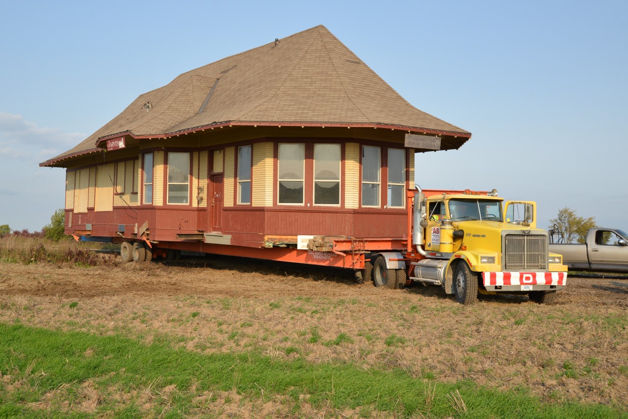The Jarvis Railway Station, a local landmark, finally embarked on its second leg of the 2 mile+ junket departing from its century acreage at the north end of the village of Jarvis at approximately 9:30 hours Tuesday, Oct 18th.  The unique ‘union station’ with its two operator bays is to be re-located to the grounds owned by the Walpole Antique Farm Machinery Association (WAFMA) at the south end of town HWY 6.  However to get this 40 feet 8” widest point x 30 feet high x 45 feet long structure to the relocation site, it had to be hauled a short distance east on the recently graded stone rail bed of the former CN Cayuga Subdivision (how appropriate is that?) then south across a clay field to a temporary resting spot at the intersection of HWY 3 and Regional RD 55.  Drainage ditches on both shoulders of HWY 3 must be filled and leveled in preparation for a tight, diagonal crossing at 8:30 hours on Wednesday, Oct 19th.  While overhead wires present challenges, the more critical part of the maneuver is guiding the station between  two uprights (poles) with mere inches to spare on either side ... center, level and steady as she goes. Once clear of HWY 3, the last leg should be less hectic albeit though another serious of fields / open land and one more drainage ditch ... to hopefully transpire before any further rainfall.  Also see info and photo id23995 dated  0327 2016 by Todd Steinman. The first leg actually took place on 13 04 2016 and simply involved moving the station off site to make way for another Tim Hortons coffee outlet.
In this photo, Cec ABRA Building Movers (Branchton, ON) can be seen turning south off of the firm rail bed of the former CN Cayuga Subdivision onto cropped fields. An 850 G Case long track was called on to provide pulling assistance to the Western Star on one brief occasion. The grey pickup truck (back right) is parked on the old rail bed.  Southern Ontario Railway (SOR) still utilize 1 km+ portion of the CN Cayuga Subdivision (with connecting track to Hagersville Subdivision) east of Regional RD 55 for rail car storage.  WAMFA is spear heading a community effort to save and relocate this historic structure as a museum and testament to the importance of railways to the heritage of the Jarvis area.  Donations from interested parties gratefully received.