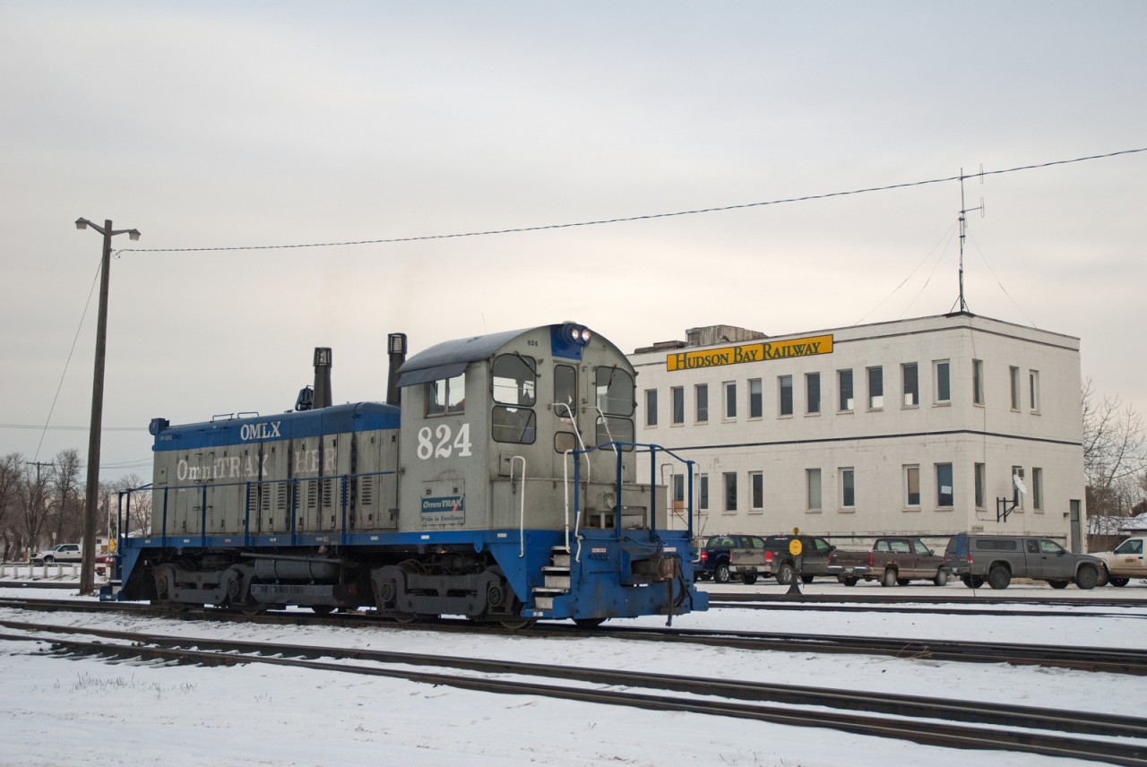 Hudson Bay Railways's SW1200 OMLX 824 pauses briefly with the company's main office visible in the background.
