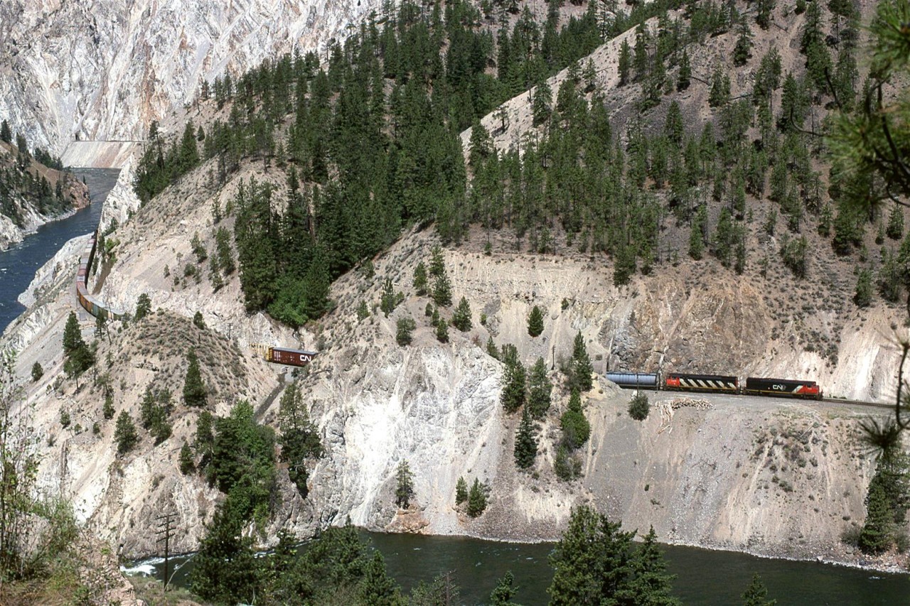 This is not the typical view of White Canyon, however, part of that view can be seen at the far left. That rocky part appears to have been severely degraded by super-heated fluids leaching out some soluble minerals. This left behind a much weakened formation. CP wisely chose the opposite river bank to build their railroad. 
The train is an eastbound manifest being led by a couple GE's.