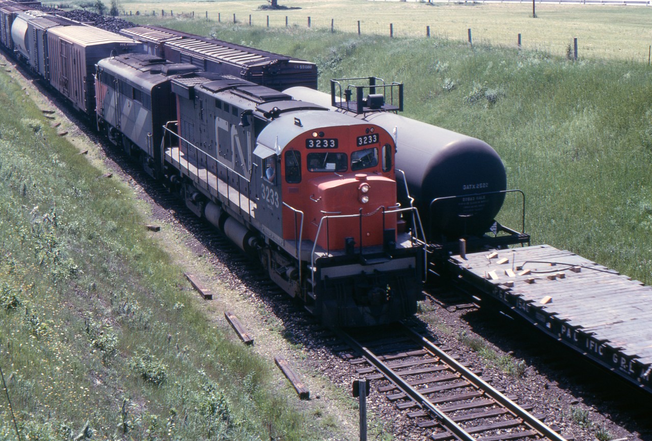 A CN eastbound with C-424 3233 and FA-2 9432 is about to pass under Waverly Road, Bowmanville on June 22, 1967. The westbound train beside it has a PGE flat car with some loose steel band lying on it. This was a hazard which railway employees working near moving trains had to watch for as it could cause serious injury.