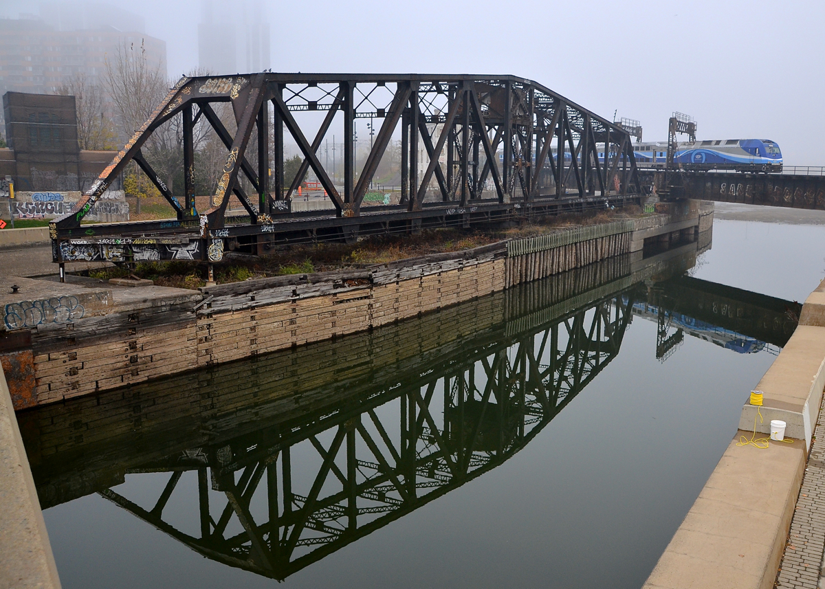 AMT 809 is arriving from the South Shore with AMT 1364 pushing it over the Lachine Canal on a foggy morning. Reflected at left is the out of use swing bridge that CN freight trains formerly used to access the Port of Montreal.