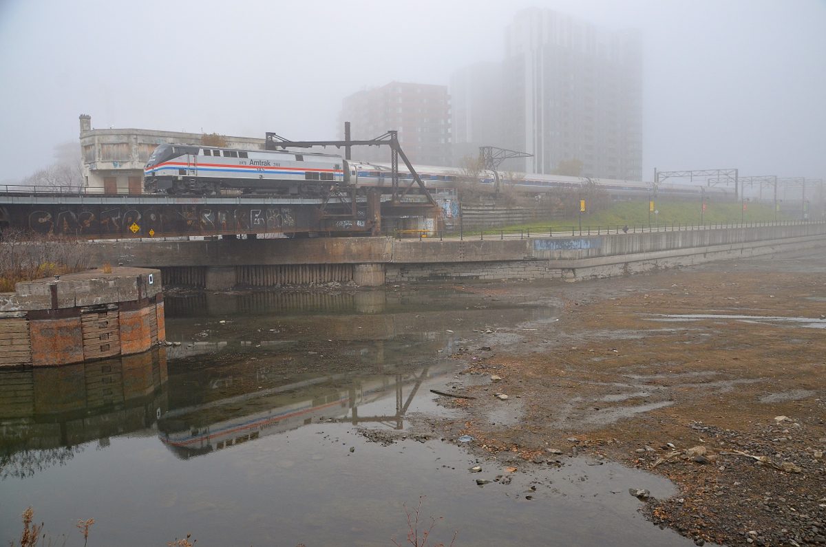 On a very foggy and windless morning, heritage unit AMTK 145 is pushing the deadheading Adirondack towards Montreal's Central Station. As happens every fall, the Peel Basin and connected Lachine Canal have been mosttly drained, but there is still enough water for the engine to be reflected.