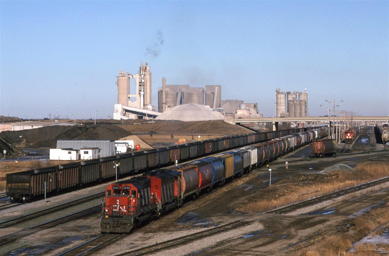 #813, a grain train for Vancouver, leaves Bissel Yard in west Edmonton. Inland Cement's plant dominates the scene.