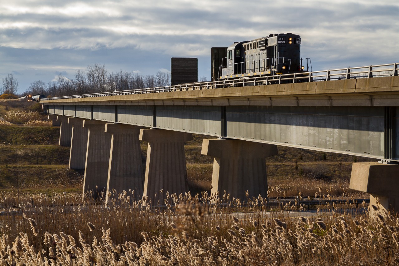 The Tunnel Bridge. At mile 4.2 of the Trillium Canal Spur, an ex-Canadian Pacific MLW RS18u guides a single car from Feeder Yard across the "Tunnel Bridge" for St, Catharines. Once operated by the Canadian National, the tunnel bridge spans a deep cut, wherein Canadian Pacific's Hamilton Subdivision runs on the approach to a tunnel underneath the Welland Canal. During the early 1970s, the Niagara Region saw a major reconfiguration of its railroad lines with the advent of the Welland Canal bypass project. On this sunny December morning, TRRY 1859 is seen working its way towards Welland and ultimately St. Catharines, while the 108 spots cars at the Atlantic Biodiesel facility in the background. Later in the day, the 108 would also cross the Tunnel Bridge to access the small Trillium yard in Welland. Once upon a time, Canadian National and Norfolk Southern shared an intermodal facility on the 108 side of the Tunnel Bridge in Dain City. Like so much of the infrastructure in the area, though, it is all but a memory.