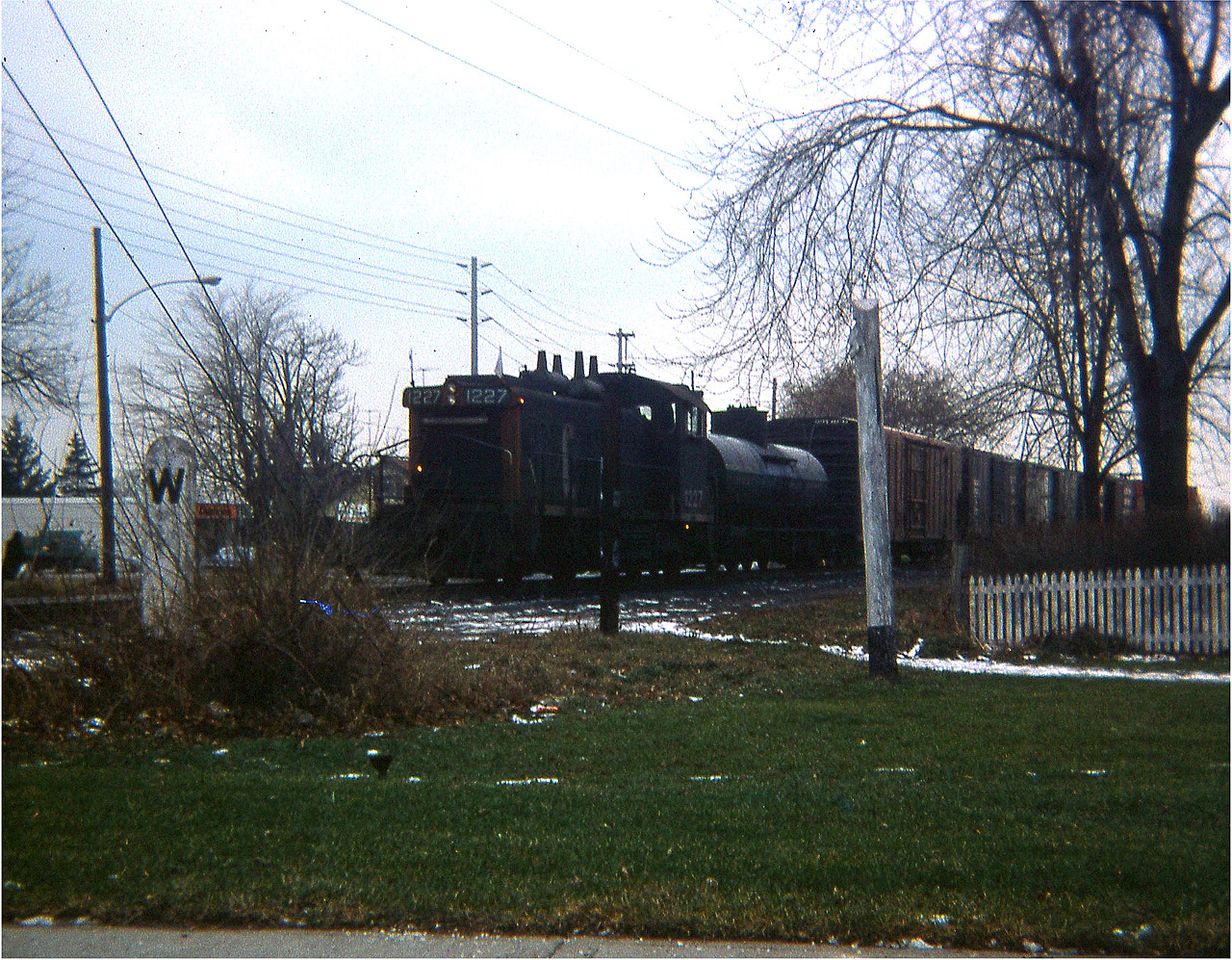 A long time back I had a really lousy little camera, a square shooter no less, and the very worst was to take a lousy camera out on a lousy day and see something good. And this happened. I had lost my old "Hawkeye" a couple years previous and still had not saved for a replacement of decent quality. So this image is the best I could do this day, despite one of the dedicated members of this group enhancing the image you see. I consider this a rare shot. CN 1227, one of the fleet of SW1200RS units, is seen pulling a lone tank and a string of boxcars up from the commercial establishment down by todays' Confederation Park in Stoney Creek. The Beach Line used to run from a connection with CN at Lake St, Stoney Creek, over the Traffic Circle, all along the beach and up to a connection with the CN main at Burlington (West) junction. When the Circle was replaced with the modern interchange at Centennial Parkway in 1969-70; the line became just an industrial run to what I remember as Langs Frozen Foods; the building still exists on the north side of the QEW between the Pwy and Burlington St exit.
In this view, taken from Elgin St looking toward Lakeshore Rd, one can see Brock St in the background, the business structure still there today. This track is long gone, and is a parking lot for an apartment building since constructed just off to the right in the photo. The line used to be busy, a passenger/freight station existed in close proximity around 75 years ago. The fact trains ran thru here once is almost all forgotten. (I wonder what happened to that nice concrete Whistle sign?)