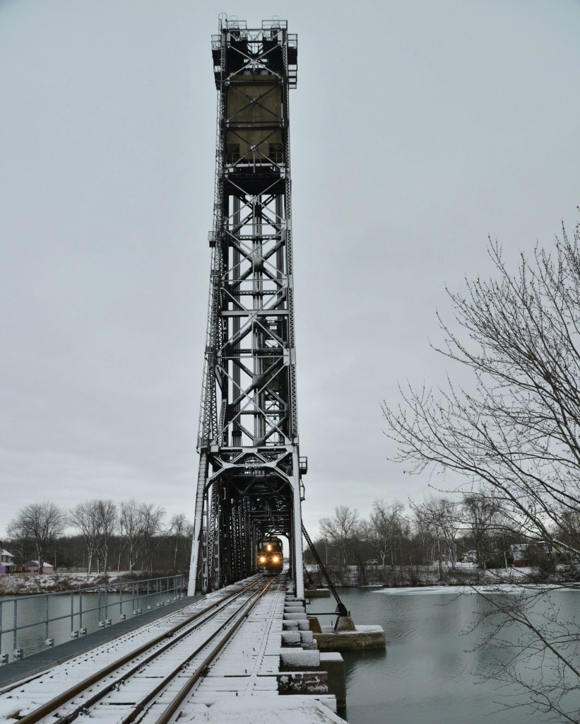 On this dull, winter's morning, TRRY 1859 trundles westbound, light power, through the massive archway of metal deck girders of Dain City's (formerly Welland Junction) 70 meter (230 feet) high twin-tower rail lift bridge. This narrow iconic 'vertical lift' rail bridge was constructed in 1927 for CNR and near identical to the nearby wider 1930 vehicular traffic bridge (Forks Road). Both bridges cross the abandoned section of the Welland Canal, a waterway in the Niagara Region,,connecting Lake Erie & Lake Ontario. No vertical lift capabilities were required for either bridge after 1980 and the towers of the vehicular bridge were subsequently removed. This old canal, renamed the Welland Recreational Waterway, hosts international rowing regattas and dragon boat races annually, and also the South Niagara Rowing Club.

This side view photo looking east hopefully depicts both the height and narrowness of this intriguing rail bridge used exclusively these day by Trillium Railway.  Craig Allen's masterpiece id= 24127 taken approximately five years prior captures its width and overall magnitude from Forks Road overlooking the stillness of the peaceful waterway. Mr. Allen picked a beautiful day to visit Dain City and was rewarded with a mixed freight of 8 cars and power to completely fill these neat, iconic structure.