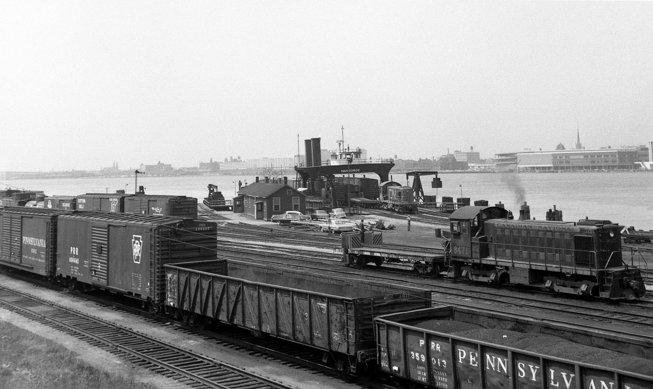 The Canadian National Windsor carferry docks and yard are shown in 1964, amid a bustle of activity: in the foreground, CN 8471 (a 1953 MLW S3 in the old switcher scheme) moves around the yard with an idler flatcar, used to keep the locomotives off the carferry during loading and unloading. In the background, N&W carferry Manitowac (built 1926 for the Wabash RR) is being loaded by another CN MLW switcher in the newer noodle scheme. In the foreground, freight cars from US roads such as the PRR, D&H and Cotton Belt congregate in the yard. And, the skyline of Detroit can be seen in the background across the river.The CN - N&W(NS) Windsor-Detroit carferry operation continued until the end of April 1994, mainly because of excess height cars that wouldn't fit in the Detroit River tunnel. When upgrades to the tunnel to handle double stack and excess height cars were complete in mid-April 1994, the carferry operations became redundant and were discontinued not long after.