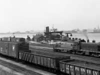 The Canadian National Windsor carferry docks and yard are shown in 1964, amid a bustle of activity: in the foreground, CN 8471 (a 1953 MLW S3 in the old switcher scheme) moves around the yard with an idler flatcar, used to keep the locomotives off the carferry during loading and unloading. In the background, N&W carferry Manitowac (built 1926 for the Wabash RR) is being loaded by another CN MLW switcher in the newer noodle scheme. In the foreground, freight cars from US roads such as the PRR, D&H and Cotton Belt congregate in the yard. And, the skyline of Detroit can be seen in the background across the river.<br><br>The CN - N&W(NS) Windsor-Detroit carferry operation continued until the end of April 1994, mainly because of excess height cars that wouldn't fit in the Detroit River tunnel. When upgrades to the tunnel to handle double stack and excess height cars were complete in mid-April 1994, the carferry operations became redundant and were discontinued not long after.