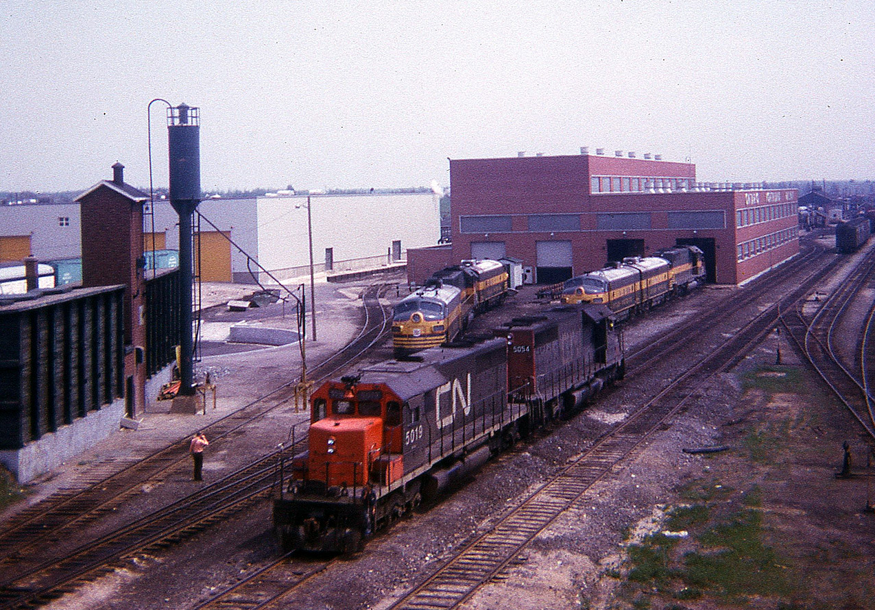 I still remember this day vividly. For a Victoria Day weekend, it was stifling hot around North Bay. In the high 80s (F), and with all the rock and pavement near the Shop, it felt like 100. Still, I am so glad I at least made a quick trek up onto the Main St overpass and captured this image at the ONR diesel facility. CN 5019 and 5054 moving around, I am guessing to get into position to take a train south to Toronto; but the fact there are four F units out front of the shop certainly indicates the age of this image. I wish I had recorded the unit numbers......oh well..........
It is worth mentioning that this sub-standard photo is the results of the cropping of an old Instamatic square slide, besides being tired by time this camera certainly wasn't the best equipment to make a trip to North Bay with, but it was better than nothing.