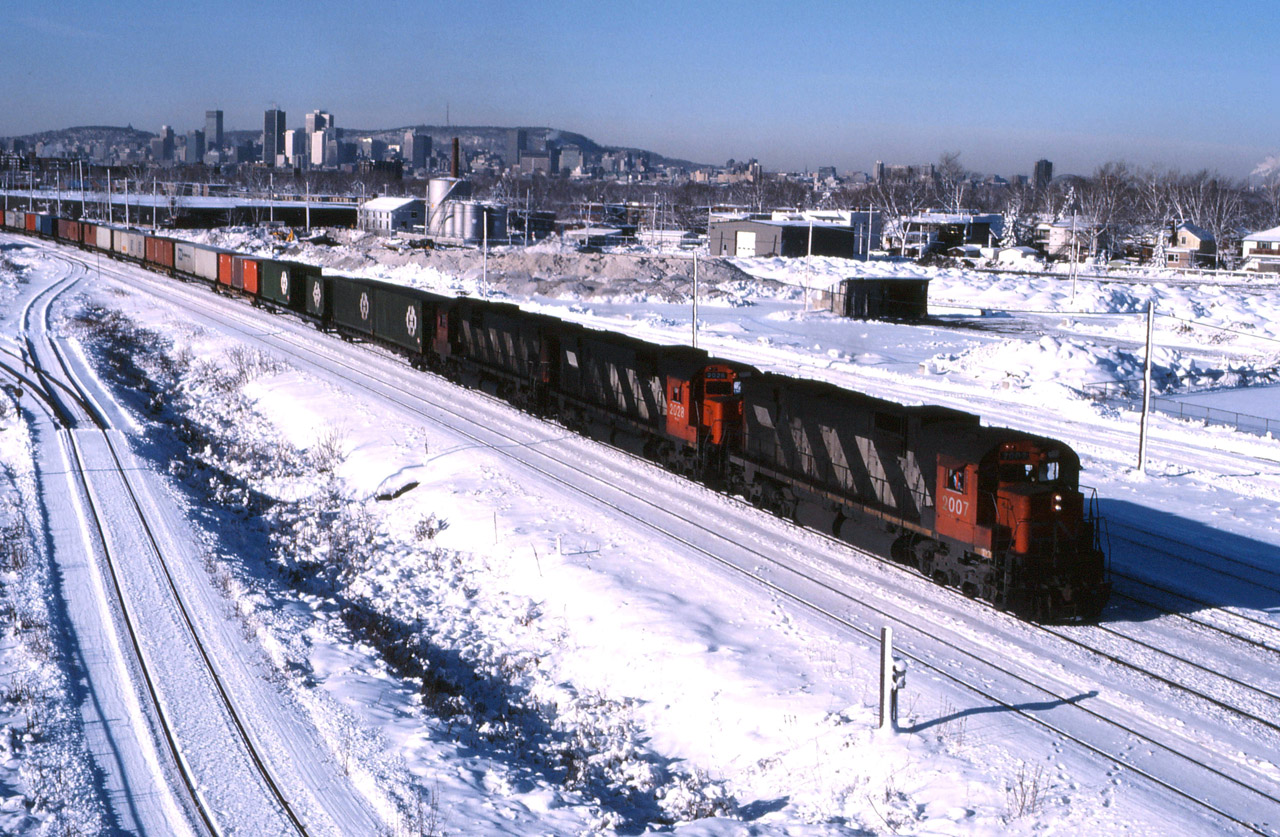 “Three C630M units and container traffic bound for the Maritimes (predominantly Halifax but some Terra Transport containers for Newfoundland are on the head end)—an eastbound heads through St-Lambert, approaching Southwark Yard. I don’t have any “hard” data on this train, but if it was siding length, the train would have had about 75-80 single level container flats cars. Compare this with the photo taken almost 30 years later at the same location (25328)—both trains have three big units (up front in 1987, DPU in 2016); about 160 containers in 1987 versus approximately 400 in 2016; in 1987, CTC extended only to St. Bruno (controlled by an operator in the St-Lambert station, then ABS/train order to St-Hyacinthe) compared to double track CTC controlled from a computerized system in downtown Montreal today; two employees up front with a third in the van versus both employees on the locomotive; CN Crown Corporation versus privatized industry leader; winter versus summer. And one photo taken by a father, the other by his son—both of whom share a passion for railroading.”