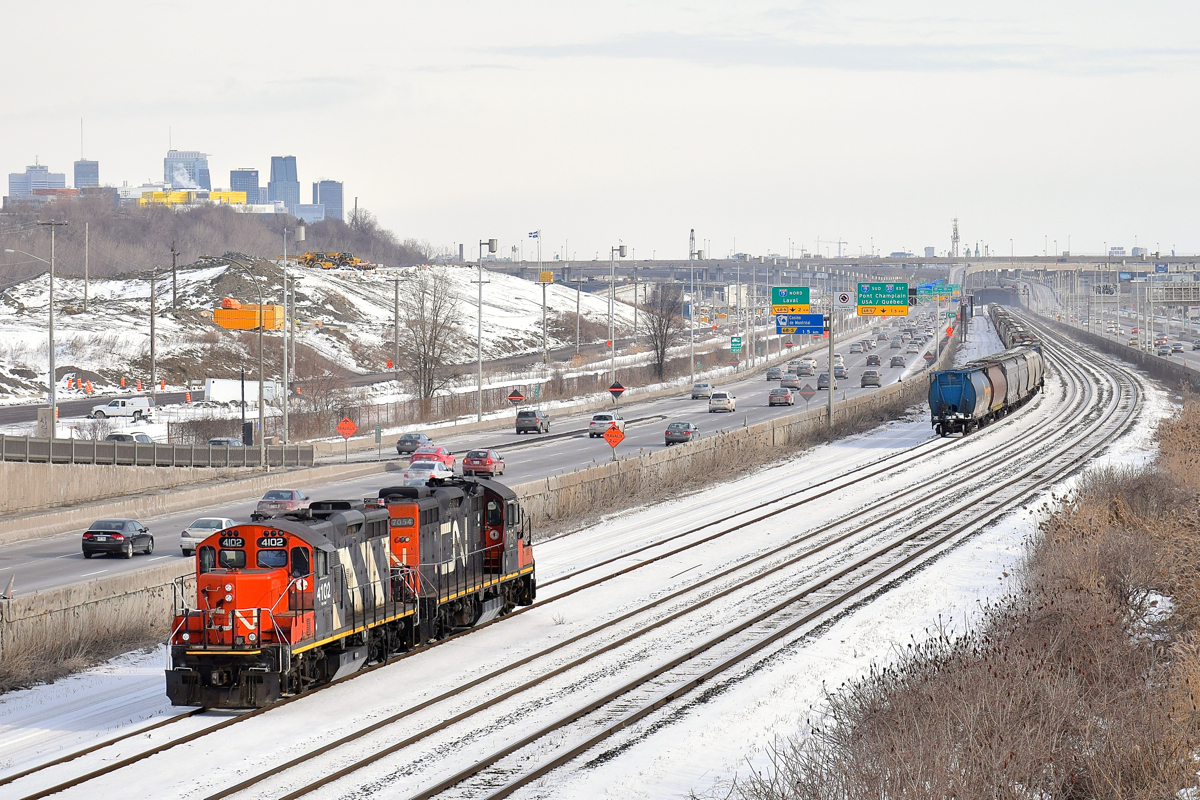 Running around. GP9's CN 4102 & CN 7054 have just  shoved 7 grain cars onto a cut of grain cars on the freight track of CN's Montreal Sub and are heading west light so that they can run around their train and head east to Southwark yard. The grain 7 grain cars had been set off by CN 874 on track 29 in Turcot Yard the day before. In the background at left is the skyline of downtown Montreal.
