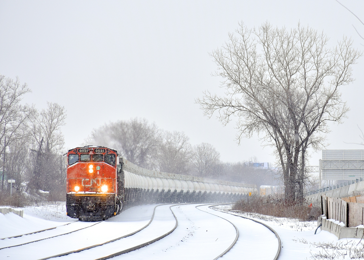 CN 585 with two strings of TankTrain loads (34 cars total) for Maitland, Ontario is westbound through Dorval on a snowy morning. Leading is GP40-2L(W) CN 9531 leading, with SD40-3 GTW 5951 trailing out of sight as the train rounds an s-curve. Barely visible in the distance is the tail end of CP 112.