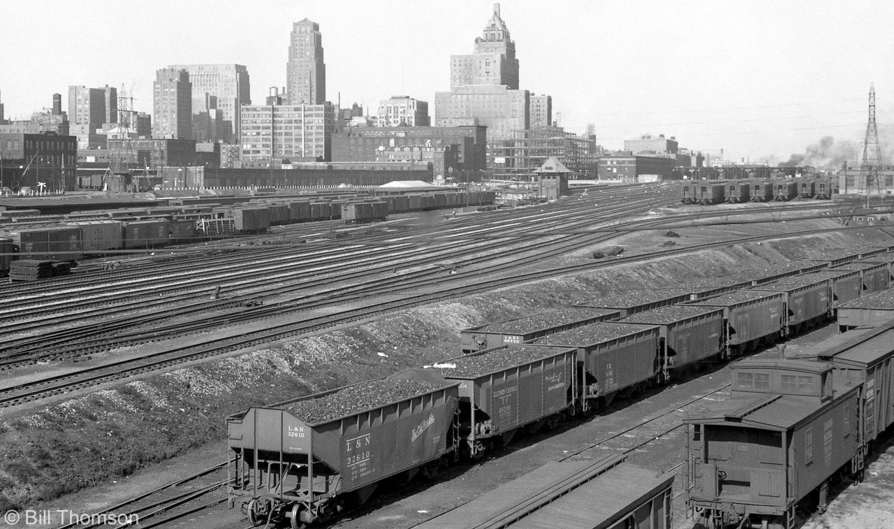 The downtown Toronto Skyline and railway corridor is pictured looking east of the Spadina Avenue overpass in 1950. 

At the bottom right, Illinois Central, Louisville and Nashville, and C&EI hoppers are spotted at CN's Spadina Roundhouse, loaded with coal for the coaling tower and steam engines that populate the ready tracks.

The passenger cars on the single-ended tracks in the upper right are in part of CPR's John St. coachyard near their roundhouse (out of view). In the distance on the upper right is Toronto Union Station and its train shed, the CN Express building, and the CN Telecommunications building under construction (later CNCP, Unitel, and presently the telecommunications hub at 151 Front St.). The main Toronto Terminals Railway tracks to/from the station pass through the middle of the image, along with the TTR's John Street interlocking tower that controlled train movements in and out of this area (one of 4 TTR interlocking towers around Union).

Toronto's 1950 skyline devoid of office towers and condos that would spring up in the decades to follow. The CPR's Royal York Hotel and the Canadian Commerce Bank Building (the tallest building in the British Commonwealth at the time) are the two most prominent buildings. Also visible on the left are CN's old freight sheds north of Front Street near Simcoe, and CN's lower freight yards with loading platforms and team tracks full of boxcars and other equipment.