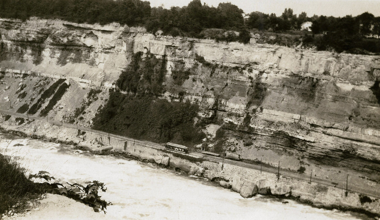 Tourists on the Great Gorge Route get a good close look at the Niagara river. A lot of rock has come down since this photo was taken by my Grandfather in 1928. The line opened in 1895 as the Niagara Falls and Lewiston Railroad, running from Lewiston to the Honeymooner Bridge near the present day Rainbow Bridge. The line was merged and connected in 1902 with the Niagara Falls Park & River Railway line on the Canadian side and became known as the Great Gorge route. Rockfalls and landslides were a constant problem. The line on the Canadian side closed in 1932. A portion of the line is visible north of the botanical gardens and butterfly conservatory. The line on the American side closed in 1935 after a 5000 ton rock slide came down just north of the whirlpool bridge. On the American side a trail runs on the roadbed from the Rainbow Bridge to below the whirlpool bridge. Location approximate, editing by the moderating team.
