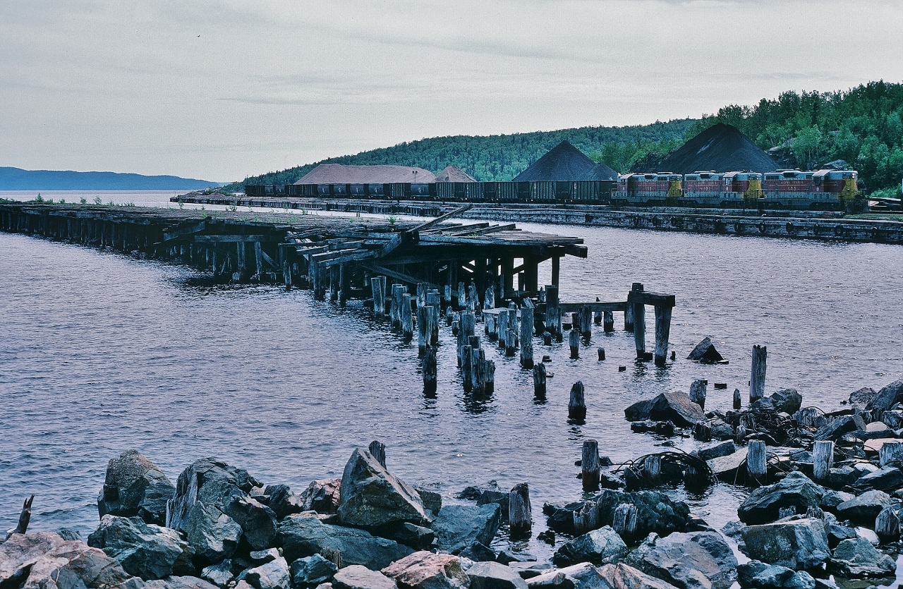 Michipicoten


...conjures the raw mysterious lands and waters of the Gitche Gumee...the ancestral and ancient lands of the Ojibway and their predecessors...


...and imagine my surprise – during the Lake Superior circle tour - after driving that Tremblay Flats Road down to the Michipicoten Harbour, to find an ACR train, with three Geeps, ready to  load up raw material for the (surprise – even at that time - operating ! ) Wawa Sinter Plant. 


ACR 103 - 101 – 102 (GMD GP7L-m rebuilt in 1978 by GMD  from ACR GP7's 155 – 169 – 165 (original GMD built 1951)), with ACR hoppers at Michipicoten Harbour  


June 25, 1985 Kodachrome by S.Danko at Michipicoten Harbour, Ontario.


P.S. For an excellent dialogue on the Michipicoten Harbour,  Sinter plant and related rail operations, see images and comment box dialogue ( & merci on this dossier to A.W. Mooney and Chris van de Heide ) :


    Sinter Plant  


    alternate dock view  


    unique ACR ore cars   


PPS: that old rail dock in the foreground demonstrates the power of Lake Superior shoreline spring ice flows. Compare the remains of the dock in this image with Arnold's 1998 image.


What's interesting: 


Sinter plants agglomerate iron ore fines (dust) with other fine materials at high temperature, to create a product that can be used in a blast furnace. The final product, a sinter, is a small, irregular nodule of iron mixed with small amounts of other minerals. (Wikipedia)


The sinter (processed iron ore & other material) from the Sinter (Wawa )plant was all shipped by rail to the Algoma Steel mill in Sault Ste. Marie. 


 Gitche Gumee ( Otchipwe-kitchi-gami - the sea of the Ojibway people )
 

sdfourty