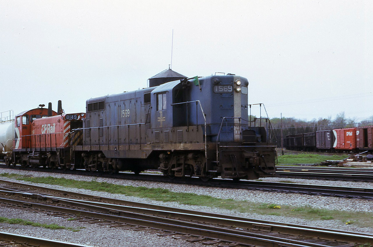 Leased Boston & Maine GP7 1569 with CP 8149 on eastbound train First 50 at Guelph Jct. in approx. April 1974. Photos of leased power displaying green flags are not all that common.  The water tower is still standing and the Goderich yard is full of cars. There are no tracks yet to store GO equipment and no enginehouse for OSR to use. The tracks in the immediate foreground are the associated connecting tracks for the Hamilton Sub. and the various yard tracks, where fans have stood for decades. Green flags, under the older Uniform Code of Operating Rules, indicated that there was (at least) another Section of this timetable train to be operated.