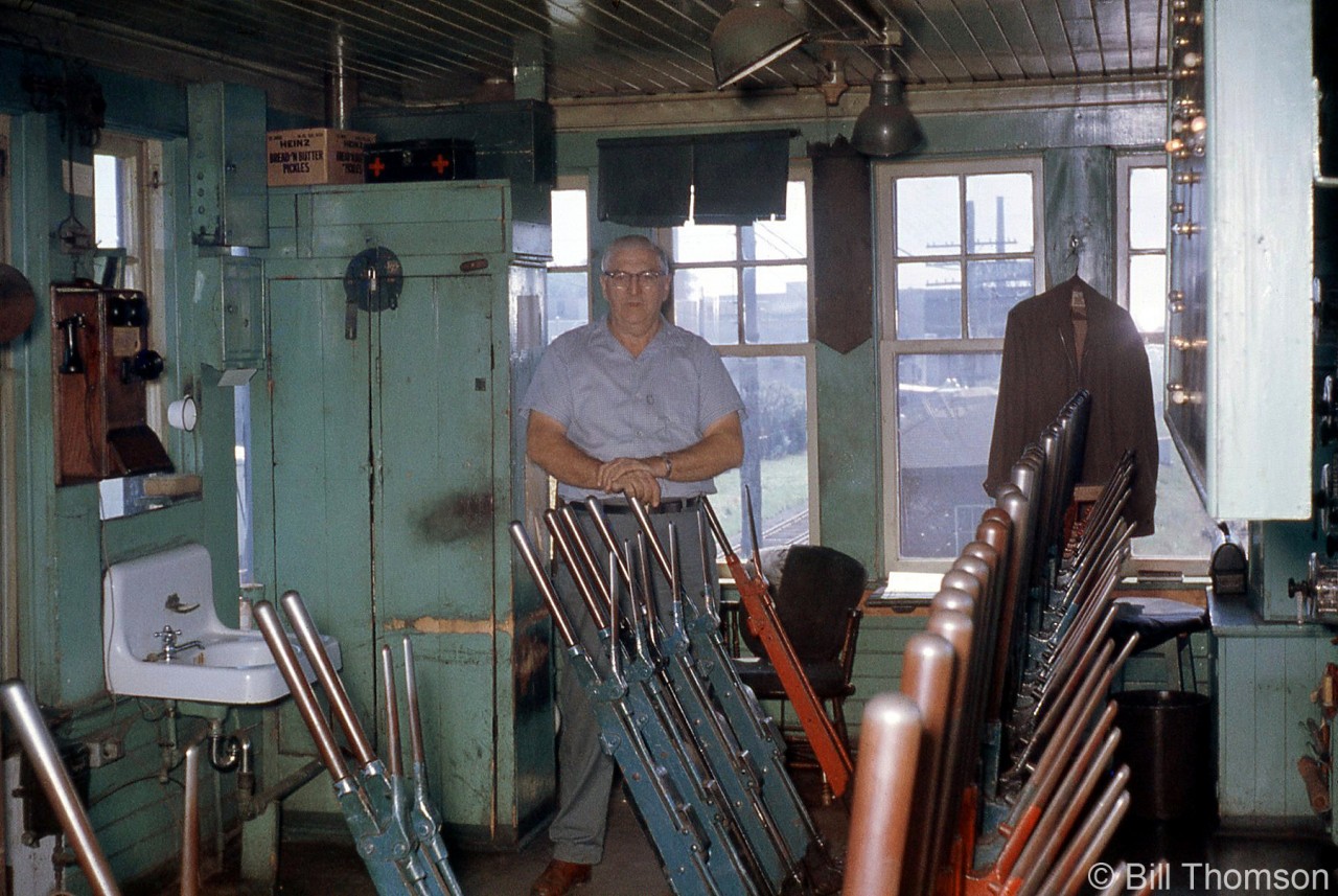 Another look inside the CP-staffed West Toronto diamond interlocking tower in February 1964, just a few months before it was demolished. The view is looking south inside, showing "leverman" Lorne Biggs next to the old Saxby & Farmer interlocking machine, its polished lever handles controlling the signals, switches, and locks in the interlocked West Toronto diamond area. The side of the illuminated track panel display is visible on the right, showing the routings and lever sequences to line specific routes.

The quaint green interior was equipped with sparse amenities, including a crank telephone, sink, storage cabinet with first aid kit (and not pictured, a stove inside for heat in the colder months). Windows all around offered good visibility of approaching trains, working track crews and rail employees in the area, and both CN's and CP's West Toronto stations were in easy view of the tower.

For more information, see the previous interior interlocking tower photo looking north-west inside: http://www.railpictures.ca/?attachment_id=28698