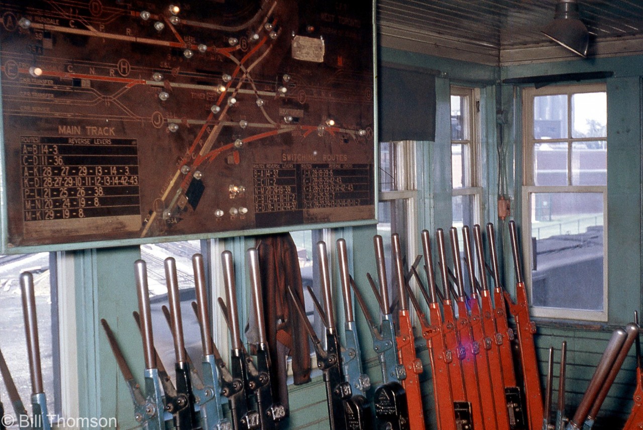 A rare look inside the West Toronto diamond interlocking tower in February 1964, not too long before it was demolished. A Saxby and Farmer interlocking machine dating from the 1900's was manually controlled by the tower's "leverman" to direct the movements of trains through "The Junction" area. The various coloured levers control track switches (blue), switch locks (black) and signals (red) through mechanical linkages of connecting rods and bell cranks that run out of the tower, along the railway lines, and to the switches and signals they control.  A large track panel display shows the various lines and routings, along with indicator lights (note the illuminated lights on the board corresponding with the CN train passing by outside). The tables of numbers on the display show the sequence of levers that must be pulled by the leverman to line specific routings.

Physically, the interlocking tower was located at the southeast corner of the West Toronto diamonds where CN's Brampton Sub (later Weston Sub) crossed CP's North Toronto Sub, and movements across both lines were directed by the tower. CP's Galt Sub (that passed by just to the west) had a connector track to the MacTier that ran northeast and bisected the diamonds. The former TG&B line (that had become an industrial spur) came off the CP MacTier Sub and ran south by the tower with its own set of diamonds, as shown on the track panel display. And, the tower also controlled movements to/from the MacTier Sub at Osler Ave. Lots of freight, passenger and local movements kept the leverman busy, along with transfer and yard movements of trains to/from CP's nearby Parkdale, Lambton, West Toronto and Agincourt Yards.

By this time, the CP-staffed and operated tower only had a few more months until modernization of the interlocking plant. According to Raymond Kennedy's Old Time Trains page, the tower was demolished in September 1964, and a shanty and switchtenders manually controlled movement over the tracks until CTC control came online in March 1965.

More at West Toronto diamond:
Exterior view of the tower and diamonds (also showing some of the control linkages): http://www.railpictures.ca/?attachment_id=16093
View of the area after demolition of the tower: http://www.railpictures.ca/?attachment_id=15874