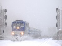 With snow continuing to fall, AMT 86 pushes some snow aside as it approaches Lasalle Station during a snowstorm. Barely visible in the background is CP's bridge over the St. Lawrence River.