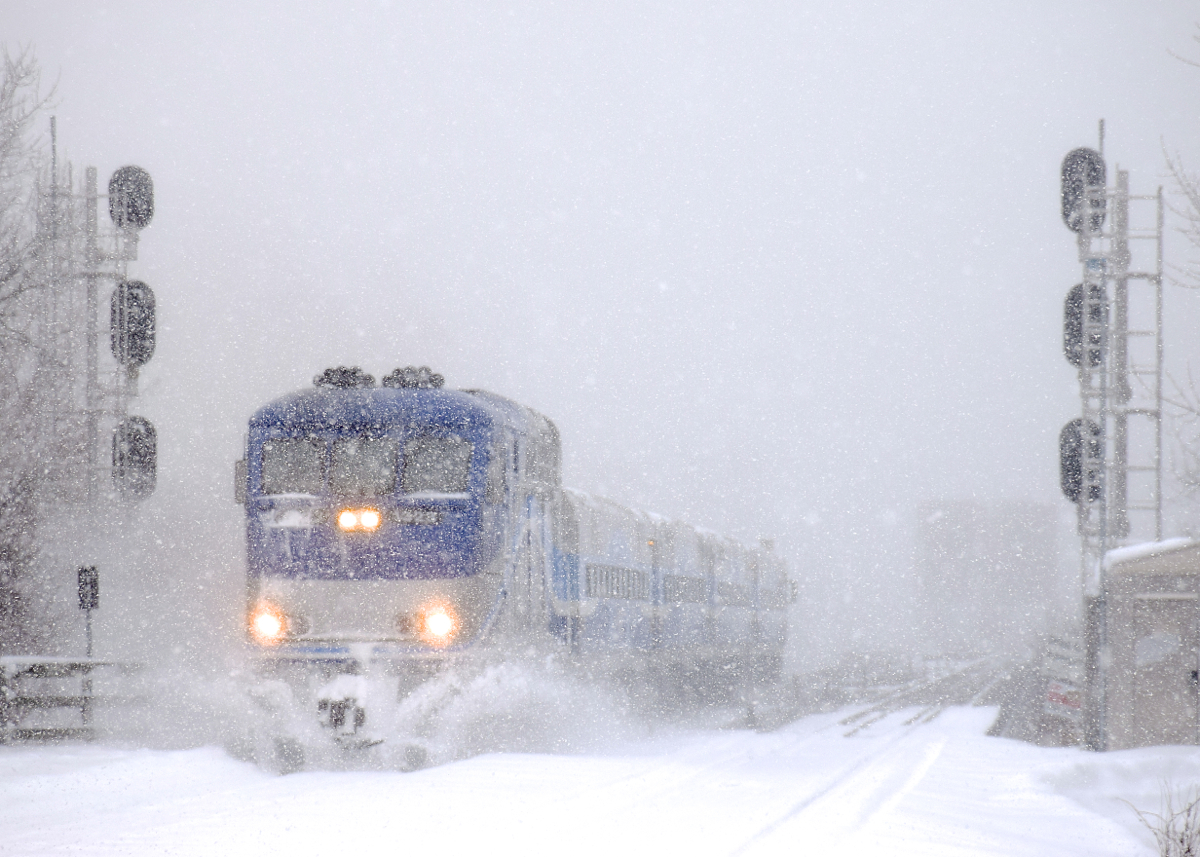 With snow continuing to fall, AMT 86 pushes some snow aside as it approaches Lasalle Station during a snowstorm. Barely visible in the background is CP's bridge over the St. Lawrence River.