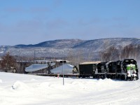 A Société du chemin de fer de la Gaspésie windmill train (which consists of 72 flat cars holding 48 windmill blades, as well as 1 loaded woodchip car up front) is climbing a grade as it rounds a curve in the small town of Maria. These windmill blades are destined for Texas.