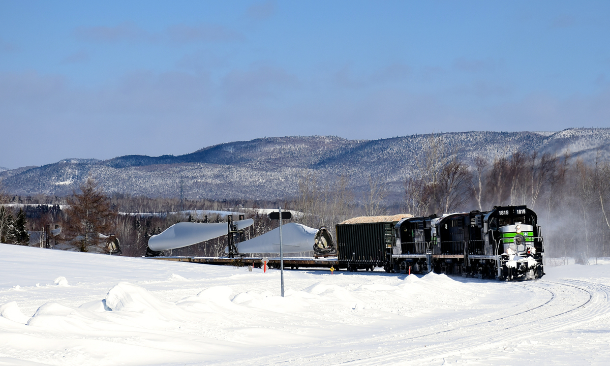 A Société du chemin de fer de la Gaspésie windmill train (which consists of 72 flat cars holding 48 windmill blades, as well as 1 loaded woodchip car up front) is climbing a grade as it rounds a curve in the small town of Maria. These windmill blades are destined for Texas.